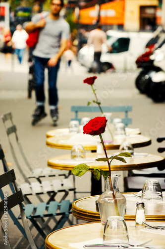 Everyday life in Paris, France. Romantic cafe terrace tables with roses in vases on sidewalk. Unrecognizable blurry young man riding roller skates at background. Selective focus on rose.