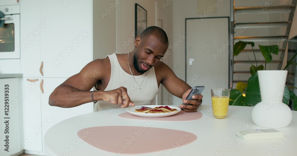 A man in pajamas eats pancakes and uses a mobile phone sitting at a table in the kitchen at home. Daylight A man in pajamas eats pancakes and uses a mobile phone sitting at a table in the kitchen at home. Daylight