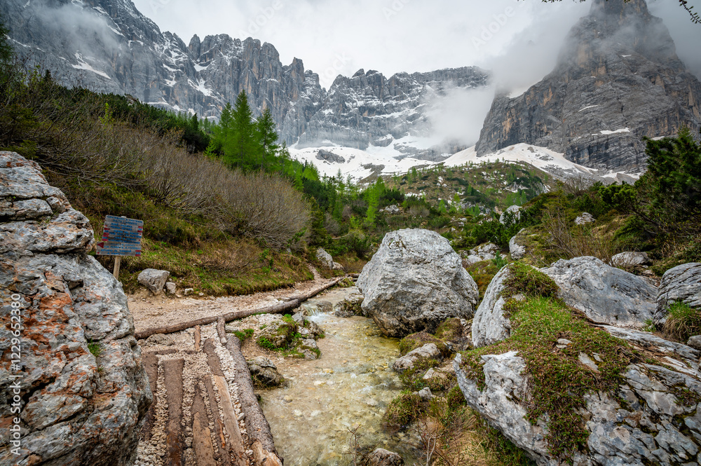 Rocky hiking trail near Lago Sorapis in the Dolomites (Dolomiti, Dolomiten), Italy. The path runs alongside a small mountain stream, with massive boulders and peaks in the background