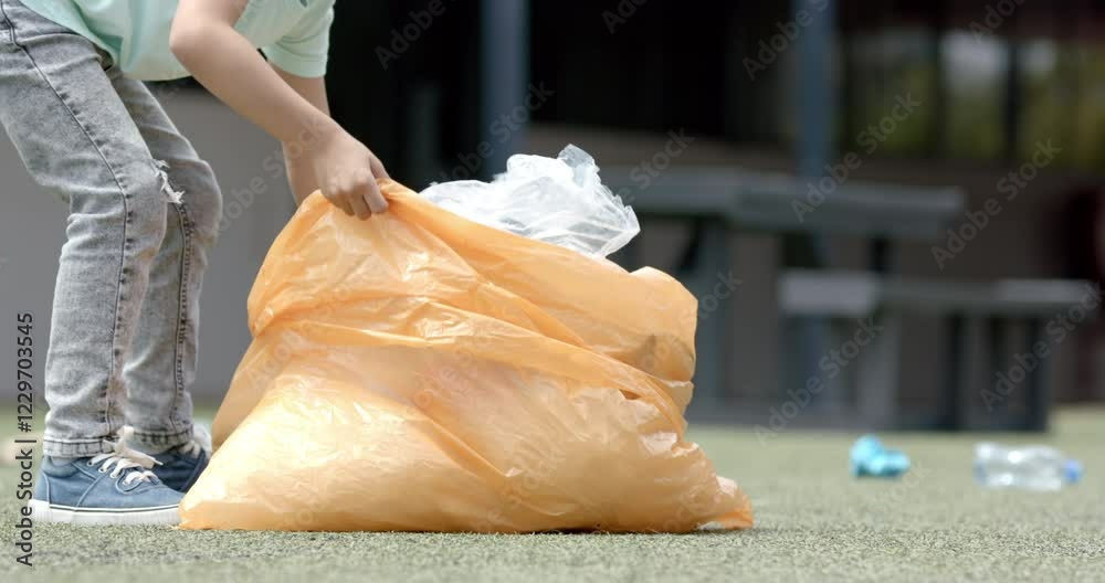 In school, child picking up trash with large orange bag, cleaning playground