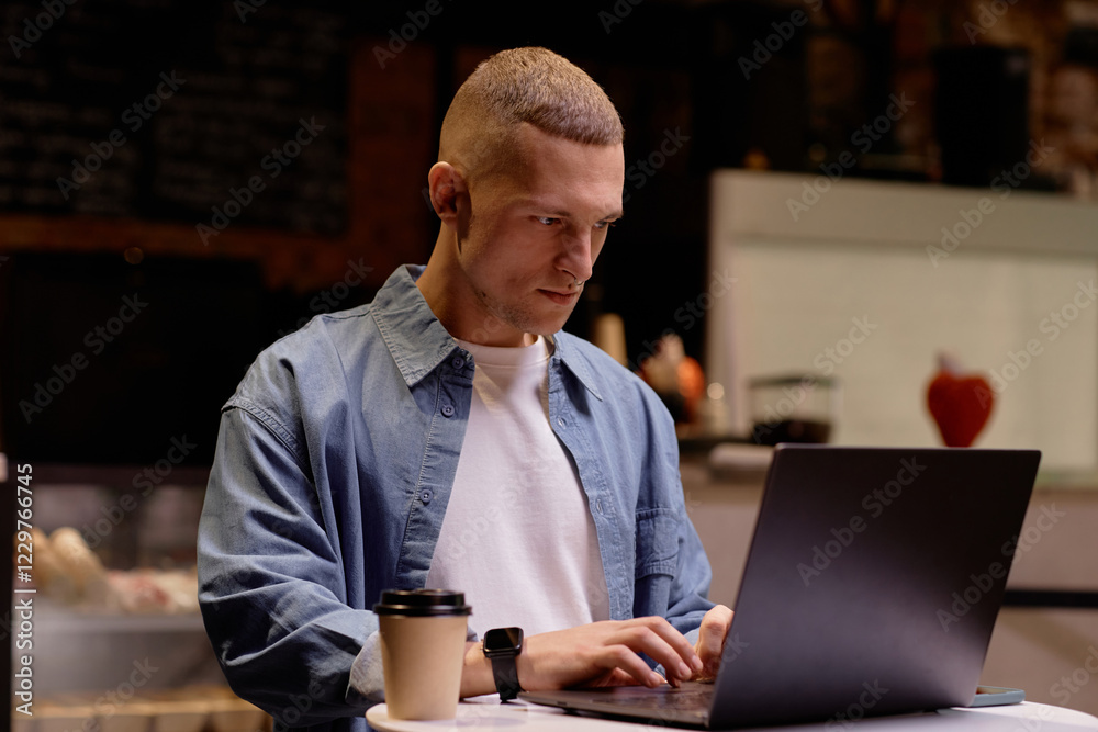 Fototapeta premium Man with short haircut working on laptop in cozy cafe surrounded by modern decor features table, coffee cup providing calm yet focused workplace ambiance