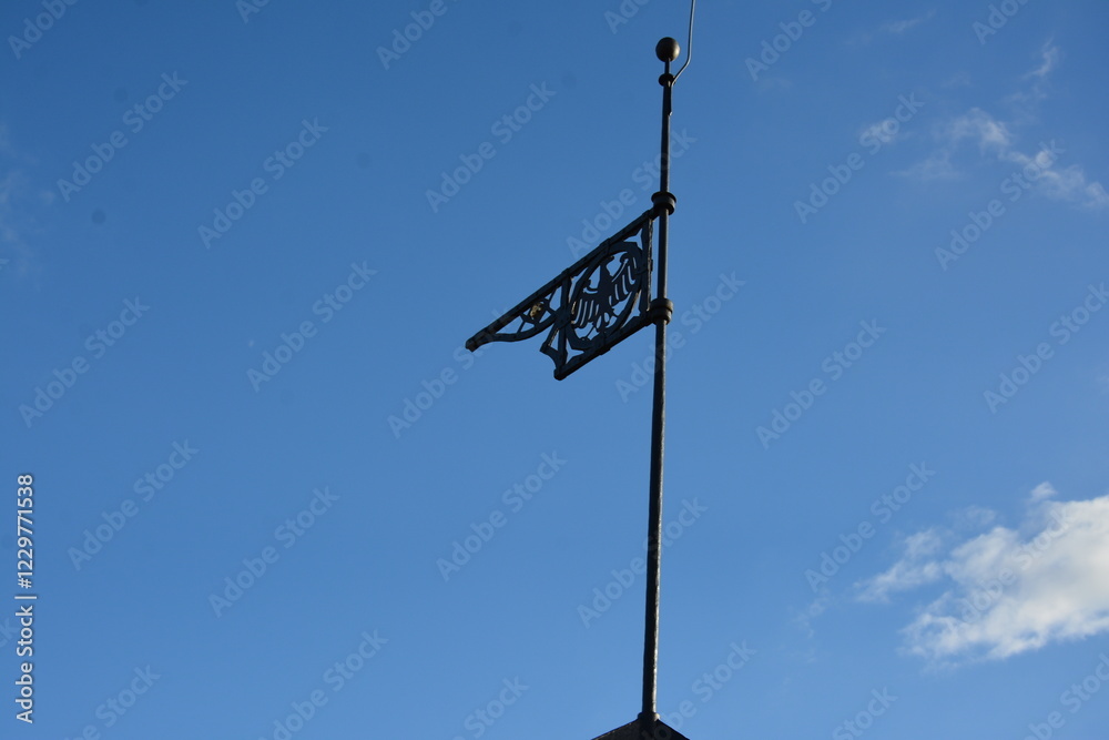 A metal weather vane with an intricate design stands atop a pole against a clear blue sky. The decorative element appears to have a pattern resembling a bird or emblem A metal weather vane with an intricate design stands atop a pole against a clear blue sky. The decorative element appears to have a pattern resembling a bird or emblem