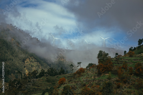 Картината върху платно A picturesque view of mountains partially obscured by fog, featuring wind turbines standing tall against a dramatic sky, showcasing nature and technology in harmony