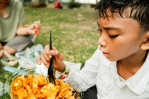 Southeast Asian boy eating tasty delicious mixed Mexican tacos tortillas chips and French fries meal in a bowl shaped plastic food container while picnic with family at park