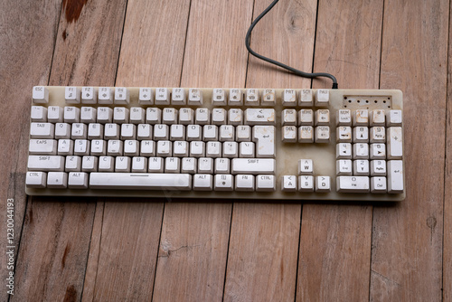 Full view of a dirty, vintage keyboard on a wooden table. Yellowed keys, visible grime, and wear. Function keys, navigation cluster, and number pad are prominent. Grey cable runs along the top.