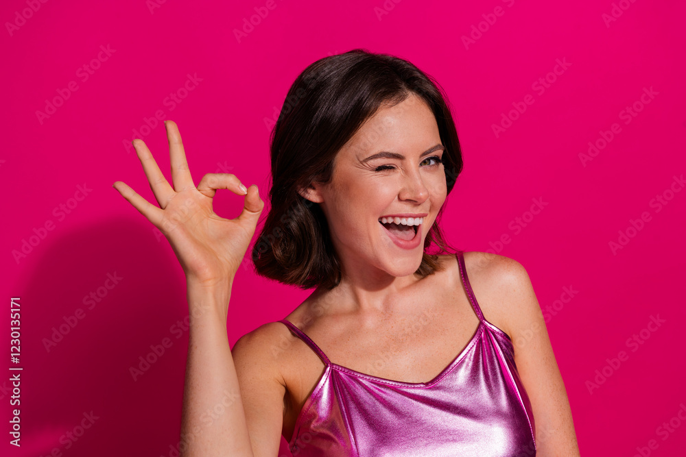 Excited young woman posing against vibrant pink background, making an OK gesture, smiling and winking Excited young woman posing against vibrant pink background, making an OK gesture, smiling and winking