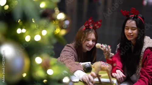 Two joyful teenage girls in festive reindeer antler headbands admire Christmas decorations and toys near a tree, enjoying the holiday season indoors with bright bokeh lights.