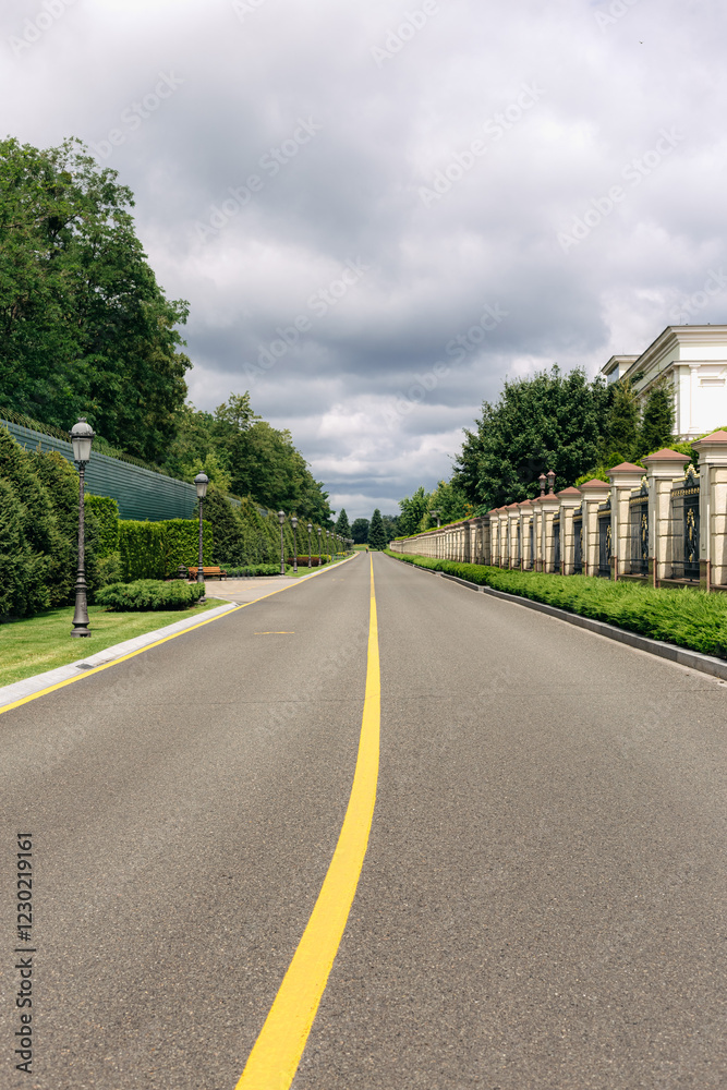 Empty asphalt road with yellow dividing strip stretching into the distance towards a cloudy sky, bordered by green trees, bushes, fences, and street lamps in a well-maintained park Empty asphalt road with yellow dividing strip stretching into the distance towards a cloudy sky, bordered by green trees, bushes, fences, and street lamps in a well-maintained park