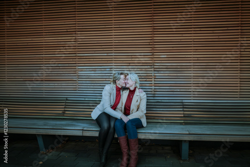 Two women sitting on a bench, sharing a loving moment, dressed in beige jackets and red tops against a wooden slatted background. photo