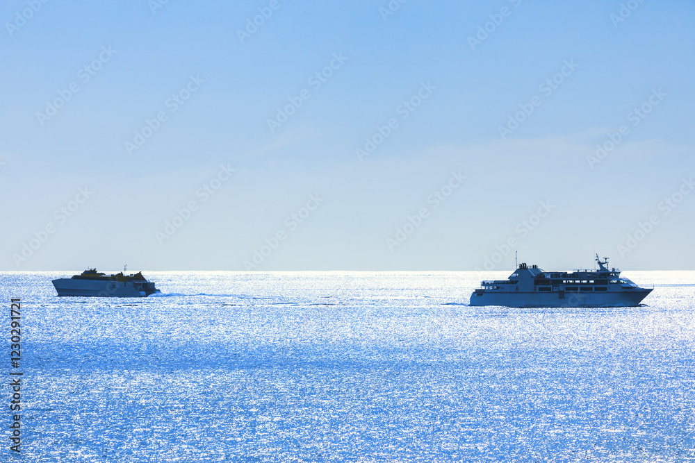 Two ships sailing on a calm, shimmering blue sea under a clear sky. Larger ship is on the right side of the image, while the smaller ship is on left, both leaving trails in water as they move forward