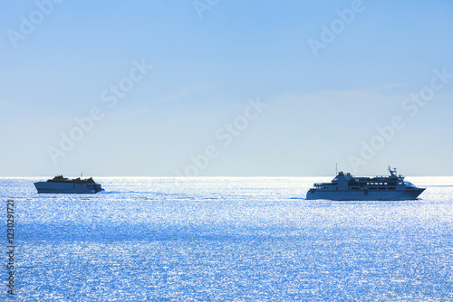 Obraz na płótnie Two ships sailing on a calm, shimmering blue sea under a clear sky. Larger ship is on the right side of the image, while the smaller ship is on left, both leaving trails in water as they move forward w salonie