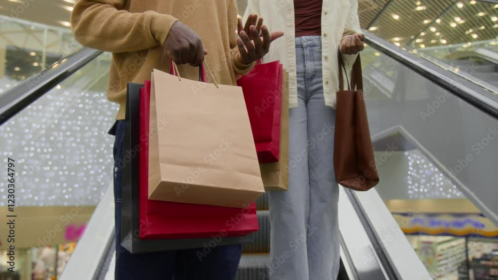 Midsection shot of anonymous friends in casual clothes holding lots of paper bags and talking while going down escalator in shopping mall, leisure time