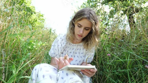 Portrait young woman in white dress sits drawing a picture in a notebook. Woman in green grass. Woman artist