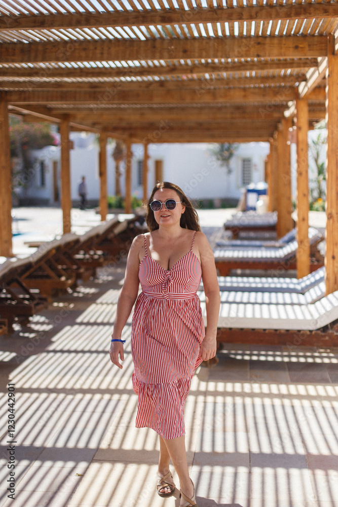 A fashionable woman leisurely strolls beneath a charming wooden pergola, delighting in the warm sun at a serene resort A fashionable woman leisurely strolls beneath a charming wooden pergola, delighting in the warm sun at a serene resort