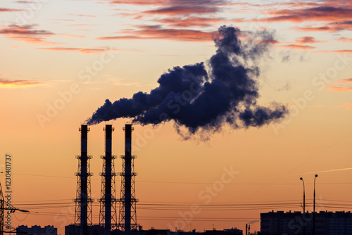 Factory chimneys release dense dark smoke into the air, colored by sunset hues. Industrial pollution of a metropolis.