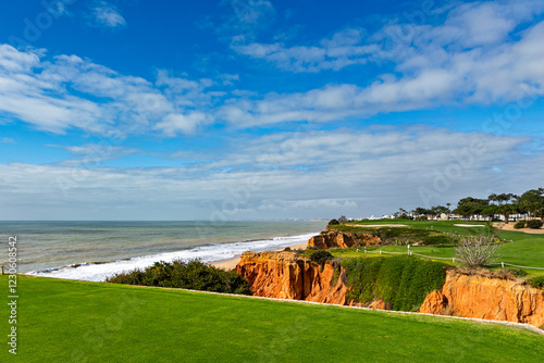 Scenic coastal golf course in Vale do Lobo, Portugal, with lush green fairways atop rugged orange cliffs overlooking the ocean. Gentle waves crash onto the shore under a partly cloudy blue sky.