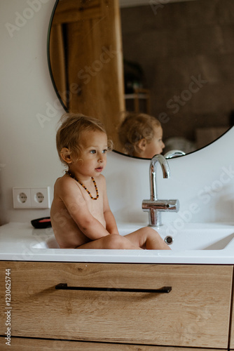 A curly-haired, beautiful and cute  little boy sits in the sink and looks thoughtful.