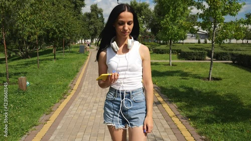 Young brunette woman in a white T-shirt listens to music in big white headphones walking along the road in a summer park. Portrait of a beautiful woman with dark hair. Woman holds a mobile phone
