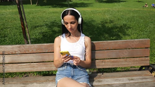 Young brunette woman in a white T-shirt and denim shorts sits on a bench in a summer park. The woman listens to music in big white headphones. Slow motion portrait woman