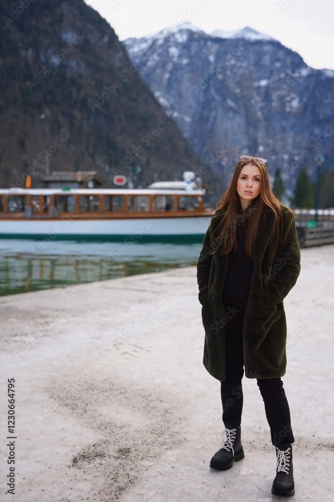 Fototapeta premium Woman at the boat station in the mountains A woman in winter clothing stands by the water, next to a boat. Snowy mountains and calm water create a peaceful and natural atmosphere.