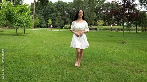 Beautiful young brunette woman in a white dress walking on the grass in a summer park. Woman with long dark hair. Slow motion