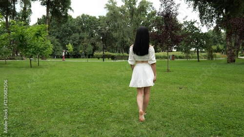 Back view of a young woman in a white dress walking in a summer park on green grass. Beautiful dark female hair. Slow motion. Beautiful summer green park
