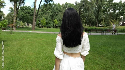 Back view of a young woman with long dark hair and a white dress. The woman is walking in a summer park. Beautiful nature around
