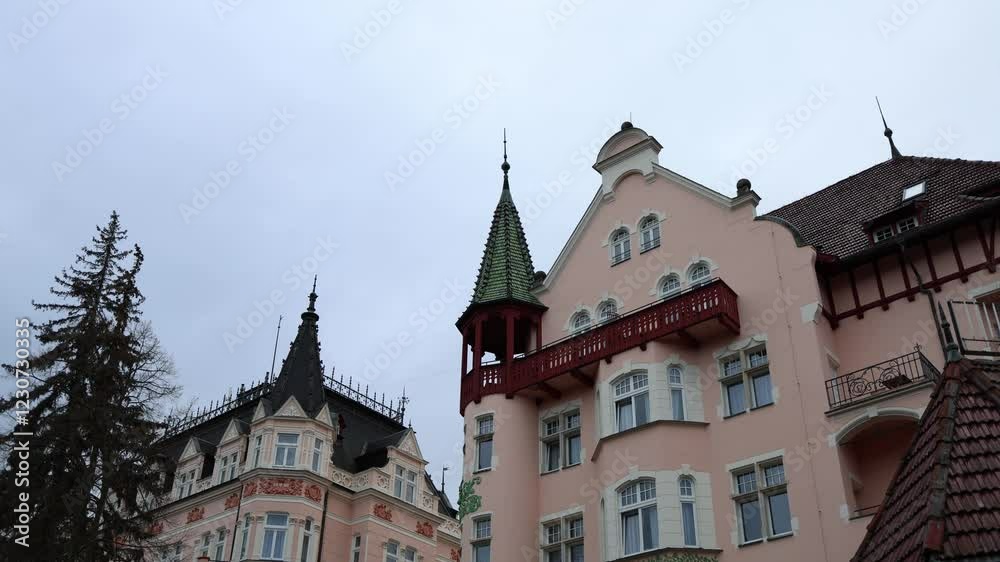 The camera tilts upwards, revealing a charming pink building with an ornate red balcony and a pointed green-roofed tower. Decorative details and contrasting roofs enhance the scene's elegance. The camera tilts upwards, revealing a charming pink building with an ornate red balcony and a pointed green-roofed tower. Decorative details and contrasting roofs enhance the scene's elegance.