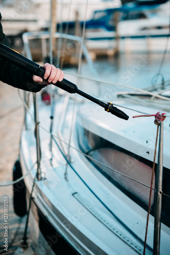 Close-up of a person using a pressure washer to clean a boat at a marina. The high-pressure nozzle is aimed at the hull, with rigging and mooring lines visible. A sharp, detailed nautical scene.
