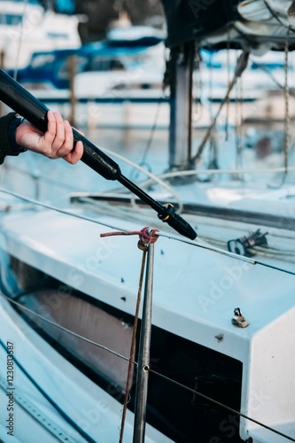 Close-up of a person using a pressure washer to clean a boat at a marina. The high-pressure nozzle is aimed at the hull, with rigging and mooring lines visible. A sharp, detailed nautical scene.
