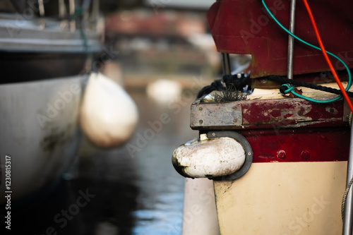 Close-up of a weathered boat stern with aged fenders and tangled ropes. The faded paint, algae growth, and mooring lines create a rustic maritime scene, with a blurred marina in the background.