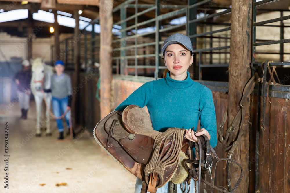 European woman horse breeder carrying leather saddle in horse barn and looking at camera. European woman horse breeder carrying leather saddle in horse barn and looking at camera.