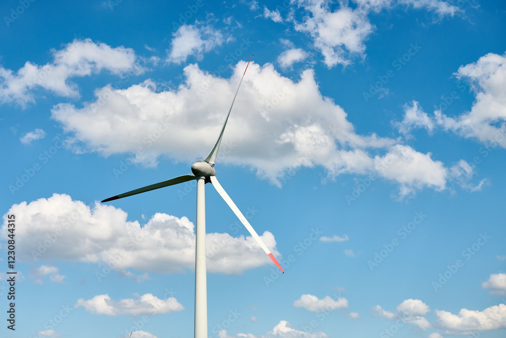 Modern wind turbine with red tipped blades stands tall against vivid blue sky filled with scattered white clouds, reflecting blend of technology and natural harmony Modern wind turbine with red tipped blades stands tall against vivid blue sky filled with scattered white clouds, reflecting blend of technology and natural harmony