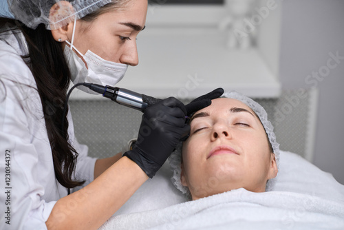 Young woman receives eyebrow permanent makeup in serene spa. Beauty technician, wearing gloves, focuses intently, creating calm, professional atmosphere