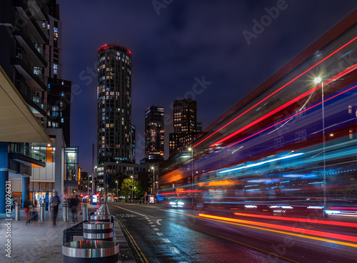 A vibrant city scene of London at night featuring tall illuminated buildings . A bus is captured in motion, creating streaks of light, blurred pedestrians.