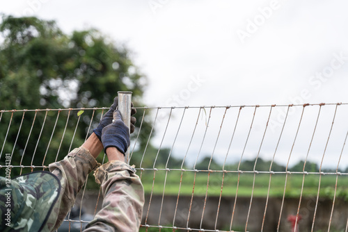 Worker in camouflage clothing secures a wire fence to a metal post outdoors, ensuring durability and stability.