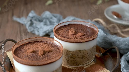 Two delicious tiramisu overnight oats in clear glass containers, layered with creamy oats, coffee, and cocoa powder, topped with chocolate pieces and coffee beans. Set on a wooden tray with a blue clo