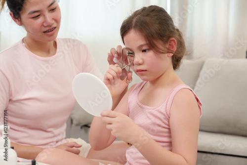 Happiness of Caucasian daughter and Asian mother as they practice eyelash curling together. Daughter carefully using an eyelash curler while mother watches supportively, creating a warm moment.