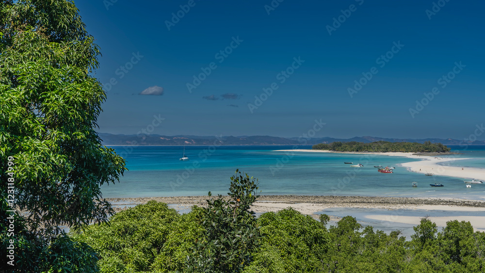 Obraz premium Beautiful postcard seascape. A sandy winding spit leads to a small green island. Silhouettes of people on the beach. Boats in the aquamarine ocean. Lush vegetation in the foreground. The blue sky.