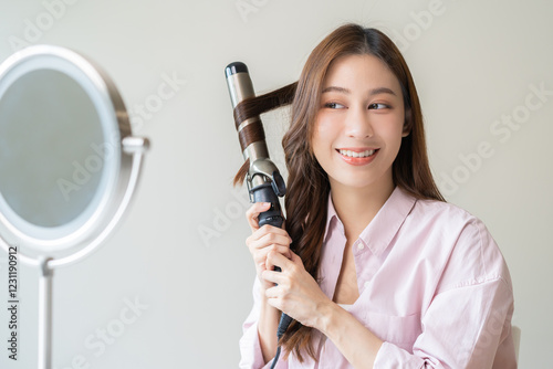 Canvas Print Beautiful curly brunette, attractive asian young woman, girl smiling using curler, curling iron long hair with ceramic curler straightener at home