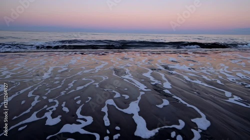 A dawn-lit shoreline where the ocean meets dark volcanic sand, with gentle surf creating swirling foam patterns.

