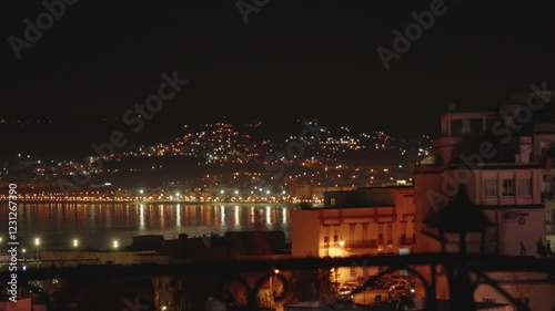 Tangier, Morocco, showcasing the cityscape with white buildings, lush greenery, and a harbor in the foreground at dusk. The hills in the background add depth to the coastal scenery
