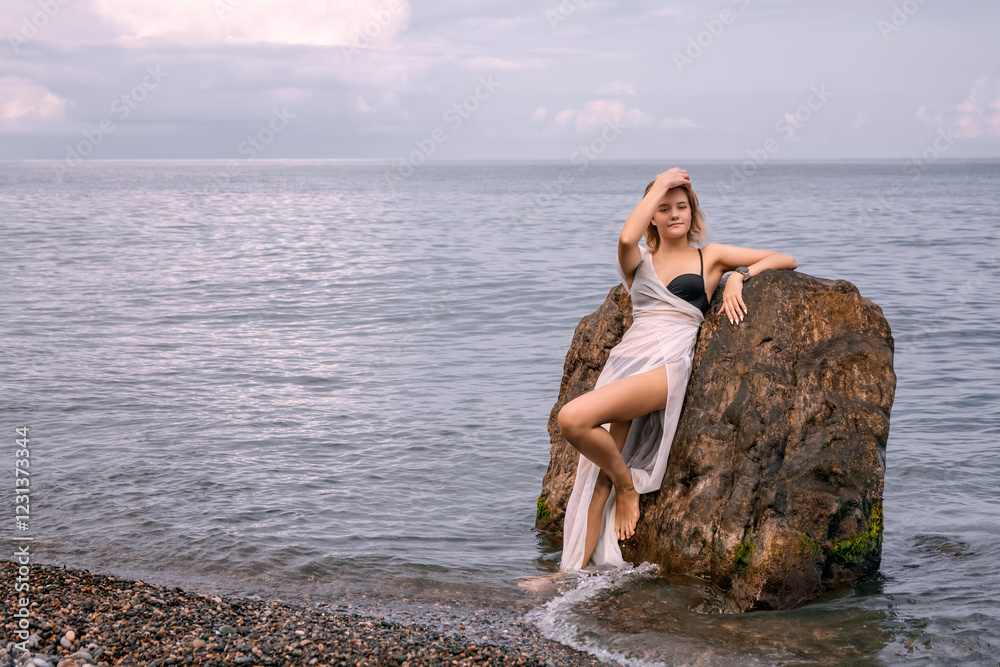 A young romantic girl in a white transparent dress stands with her back against a rock in the sea on a cloudy day. She has a smartwatch on her wrist. The concept of a modern goddess A young romantic girl in a white transparent dress stands with her back against a rock in the sea on a cloudy day. She has a smartwatch on her wrist. The concept of a modern goddess