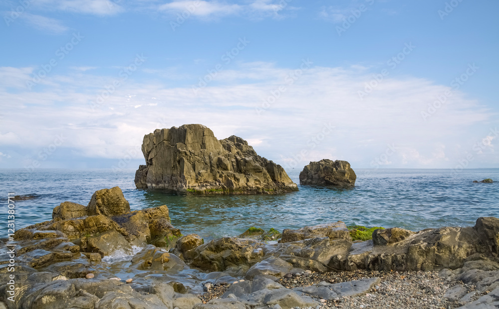 Rocky wild coast. A cliff in the sea. Blue sky with light clouds. Ripples on the clear blue water. Landscape with reefs. Tsikhisdziri Beach. Georgia. Rocky wild coast. A cliff in the sea. Blue sky with light clouds. Ripples on the clear blue water. Landscape with reefs. Tsikhisdziri Beach. Georgia.
