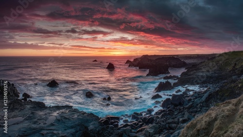 Vibrant sunset hues of orange and pink blending into dark clouds over a rugged rocky coastline with crashing waves and exposed black rocks