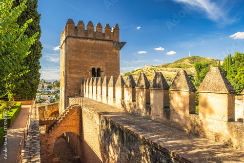 Alhambra complex in Granada town, Andalusia, Spain. Ancient fortress walls and tower overlooking a cityscape under a vibrant blue sky.
