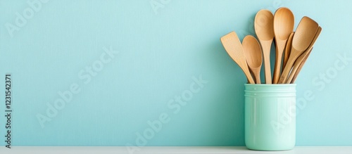 Wooden kitchen utensils in a light teal jar positioned on a white countertop against a soft pastel blue wall, creating a clean and inviting backdrop. photo