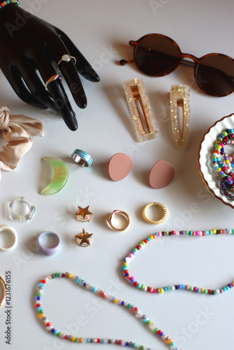 Hand figurine, sunglasses, gold jewelry, colorful bead jewelry, statement rings and various hair accessories on the white table. Selective focus.