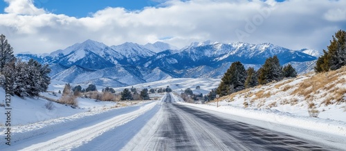 Snow-covered mountain landscape with a clear blue sky, featuring a wide snowy road stretching towards dramatic peaks and evergreen trees on the sides.