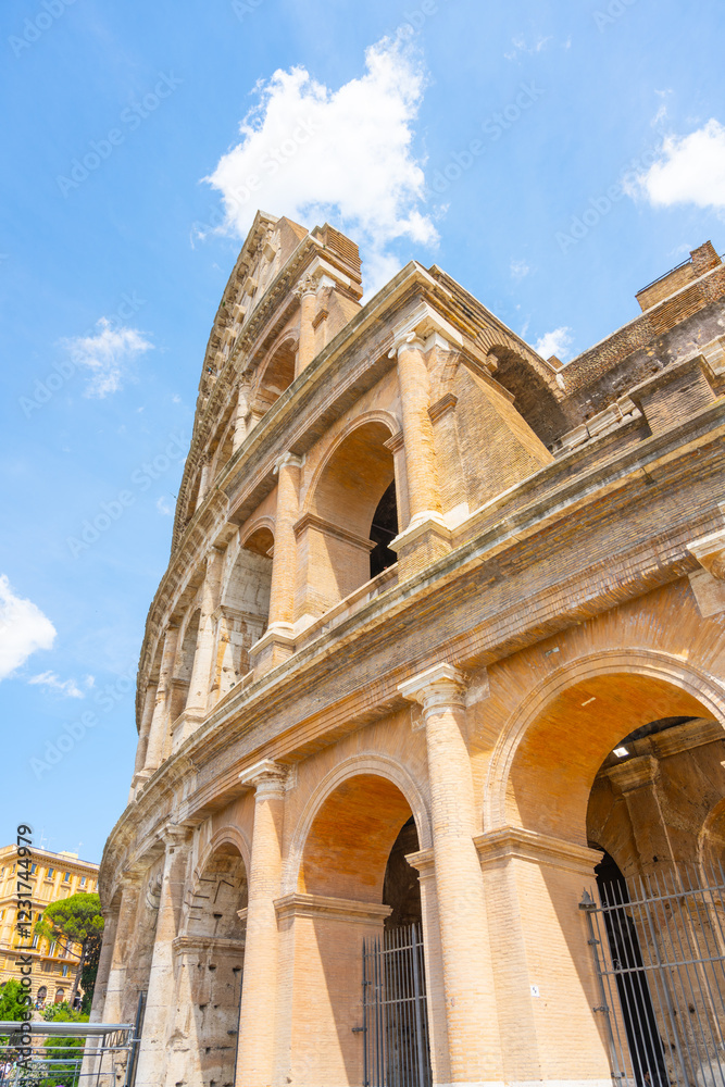 Fototapeta premium The iconic Colosseum in Rome stands tall against a bright blue sky, showcasing its ancient architecture and intricate arches. Visitors enjoy the grandeur of this historic landmark.